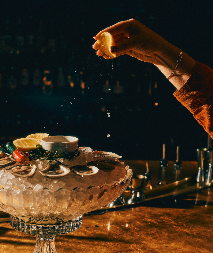 A hand squeezing lemon over a crystal pedestal dish of oysters on ice at a dimly lit bar.