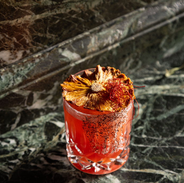 A red cocktail in a crystal rocks glass garnished with a dried flower, set on a green marble surface.