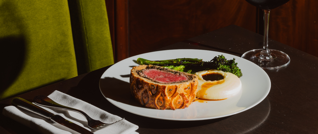 A sliced beef wellington with a pink center, served with asparagus and greens on a white plate alongside a glass of red wine.